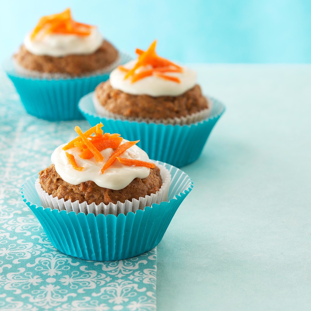 "Three cupcakes in blue wrappers topped with white frosting and orange decorative garnish, arranged on a patterned surface against a light blue background