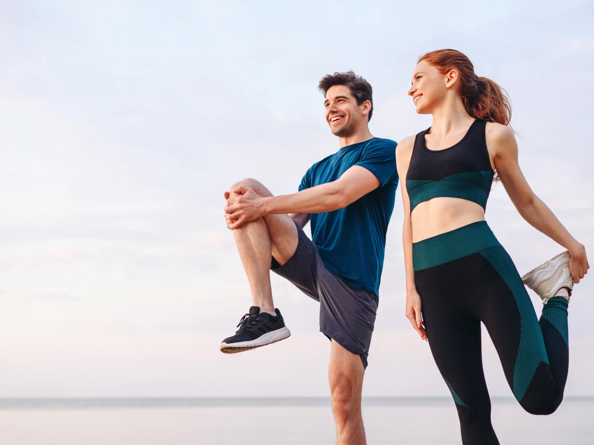Two people in athletic wear perform leg stretches, one wearing navy blue top and one wearing black top