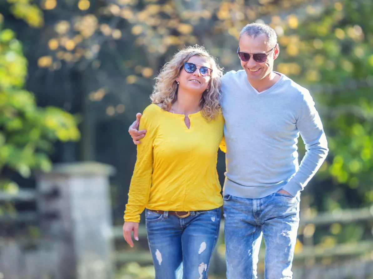 Two people walk outdoors on path, one wearing yellow top with sunglasses, one wearing light blue top, both in jeans