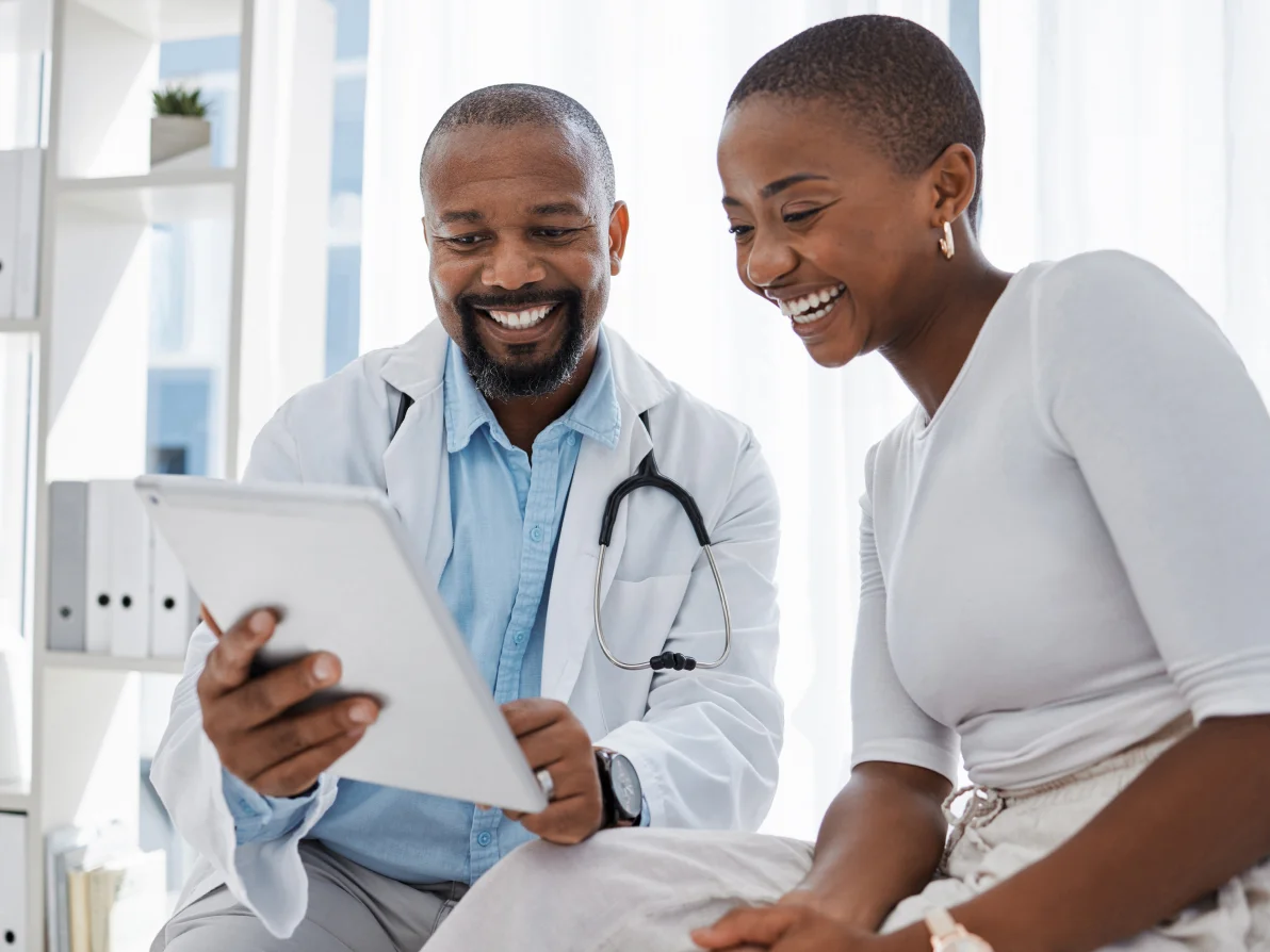 Man in light blue shirt with stethoscope and woman in white coat smile while looking at mobile device together