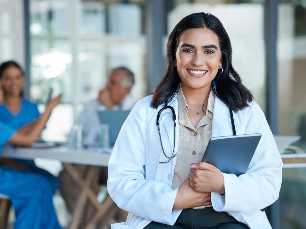 Woman with long dark hair in white medical coat and stethoscope holds blue folder or tablet in bright office