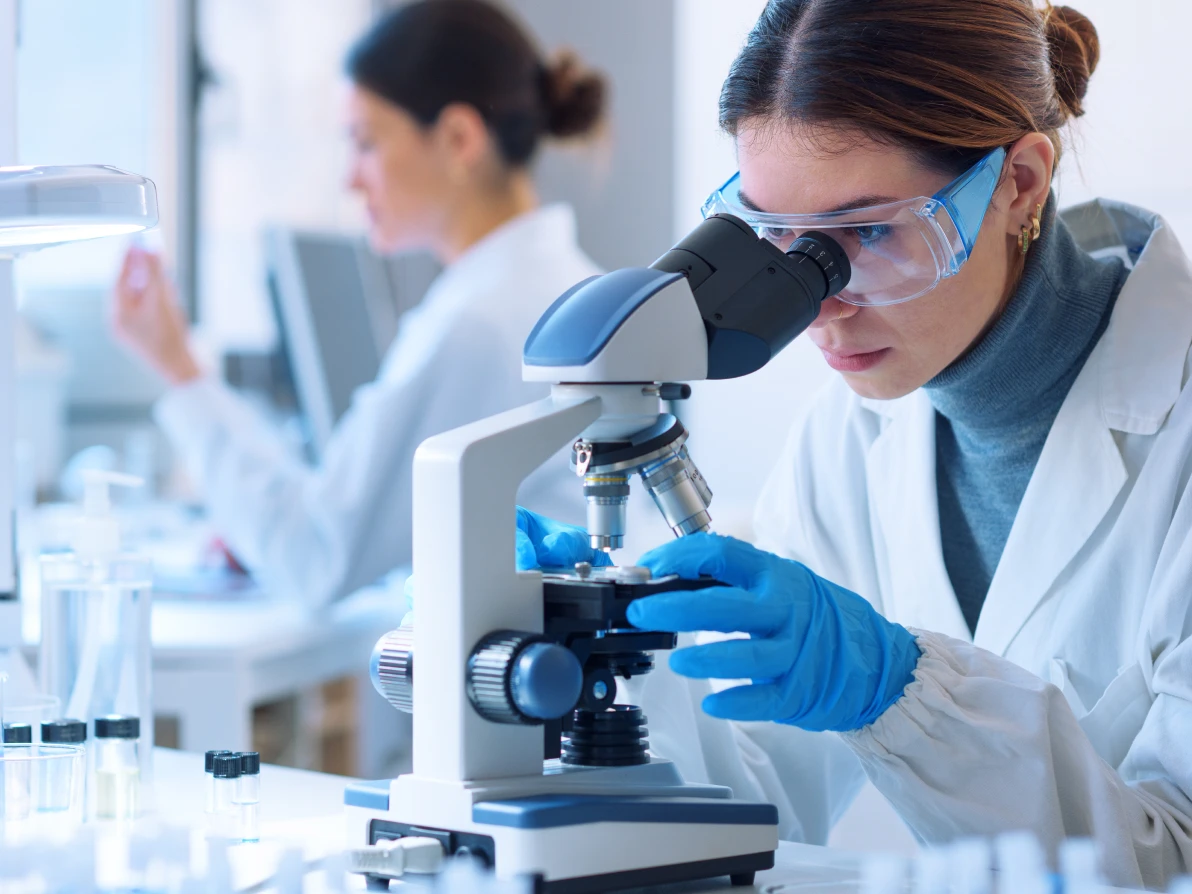 Two people in white lab coats work at microscope in laboratory, one wearing blue gloves