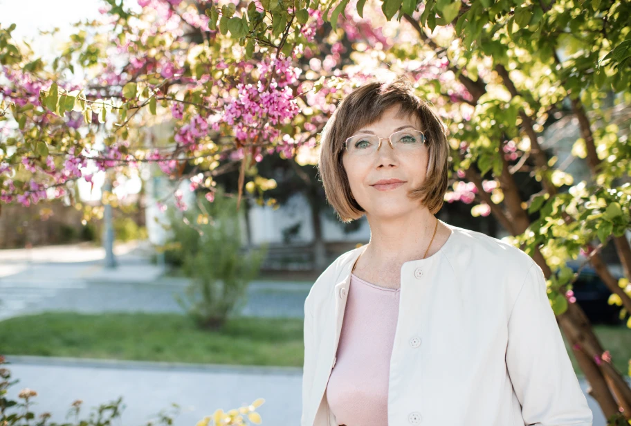 Person in white top and pink cardigan standing outdoors with flowering plants visible