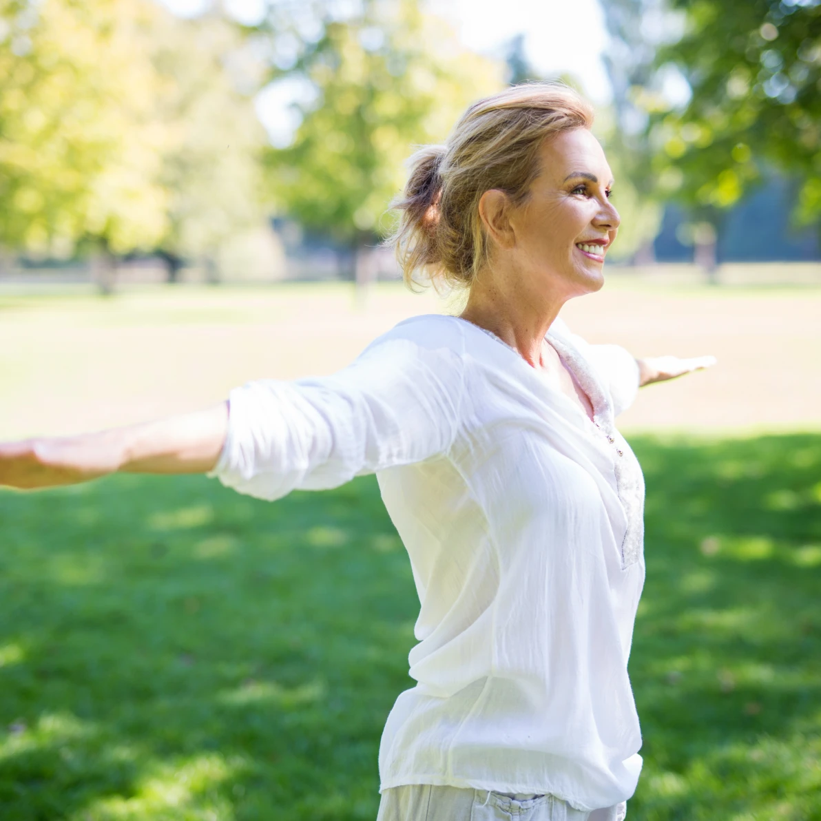 Person in white long-sleeve top with arms extended outdoors in park setting