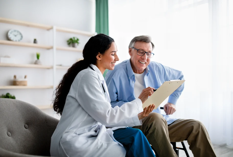 Person in white coat and person wearing glasses and blue shirt sitting together looking at a clipboard in a bright room