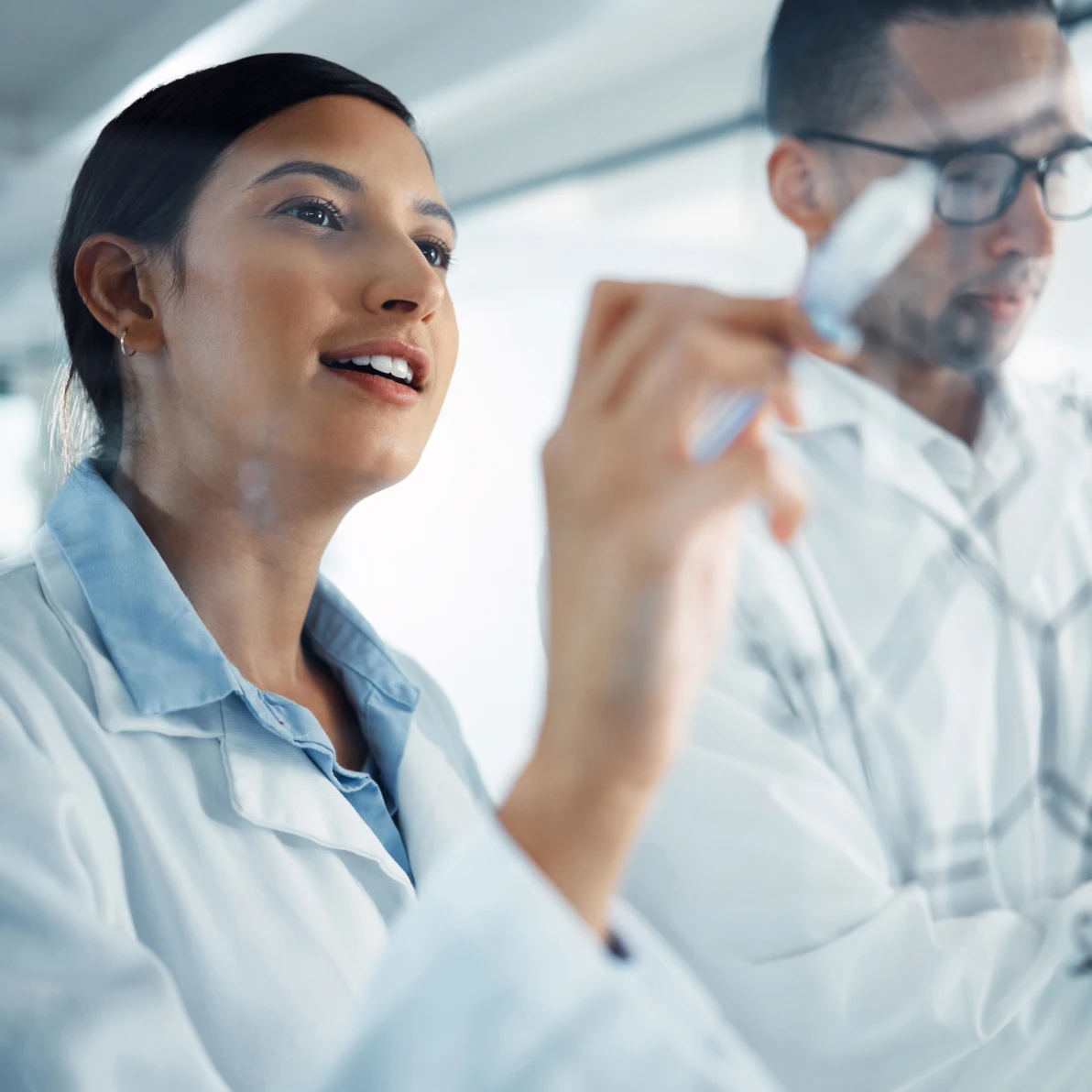 Person in light blue collared shirt gesturing toward a transparent surface in a bright office setting, another person wearing glasses visible in background
