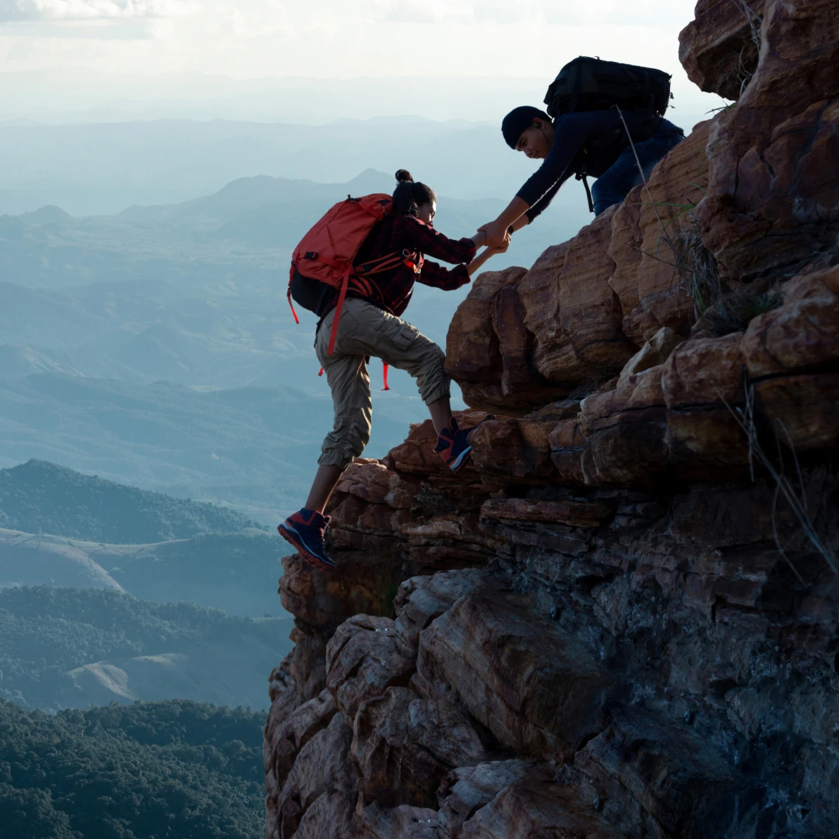 Two people climbing on rocky terrain with one person extending a hand to assist the other, mountain landscape visible in background