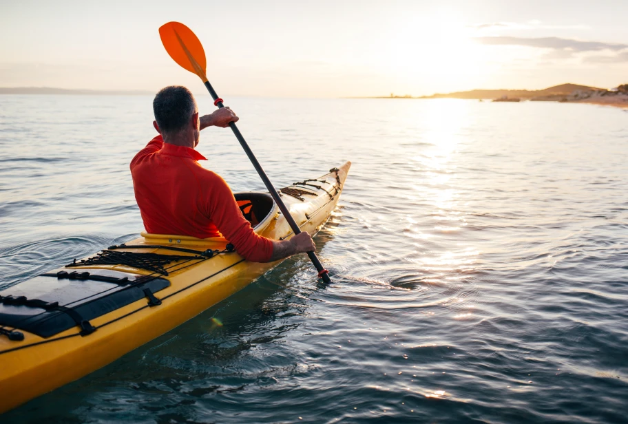 Person in red top paddles yellow kayak on calm water during golden hour