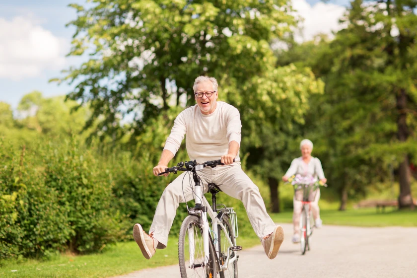 Person in light-colored top rides bicycle on tree-lined green path with others visible in background