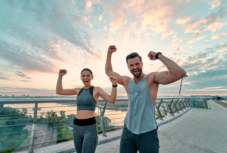 Two people in athletic wear raise arms in celebration pose at waterfront during sunset