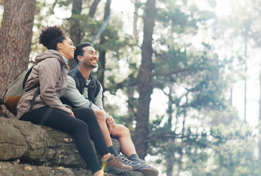Two people sit on fallen log in forest, one wearing brown jacket and one wearing gray jacket