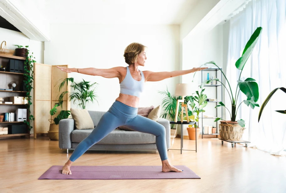 Person in white top and blue pants performs lunge position on purple yoga mat in bright room with plants