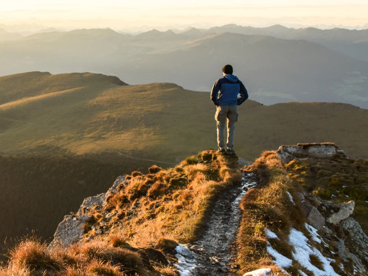 Person in dark jacket stands on rocky mountain peak overlooking valley at sunset or sunrise