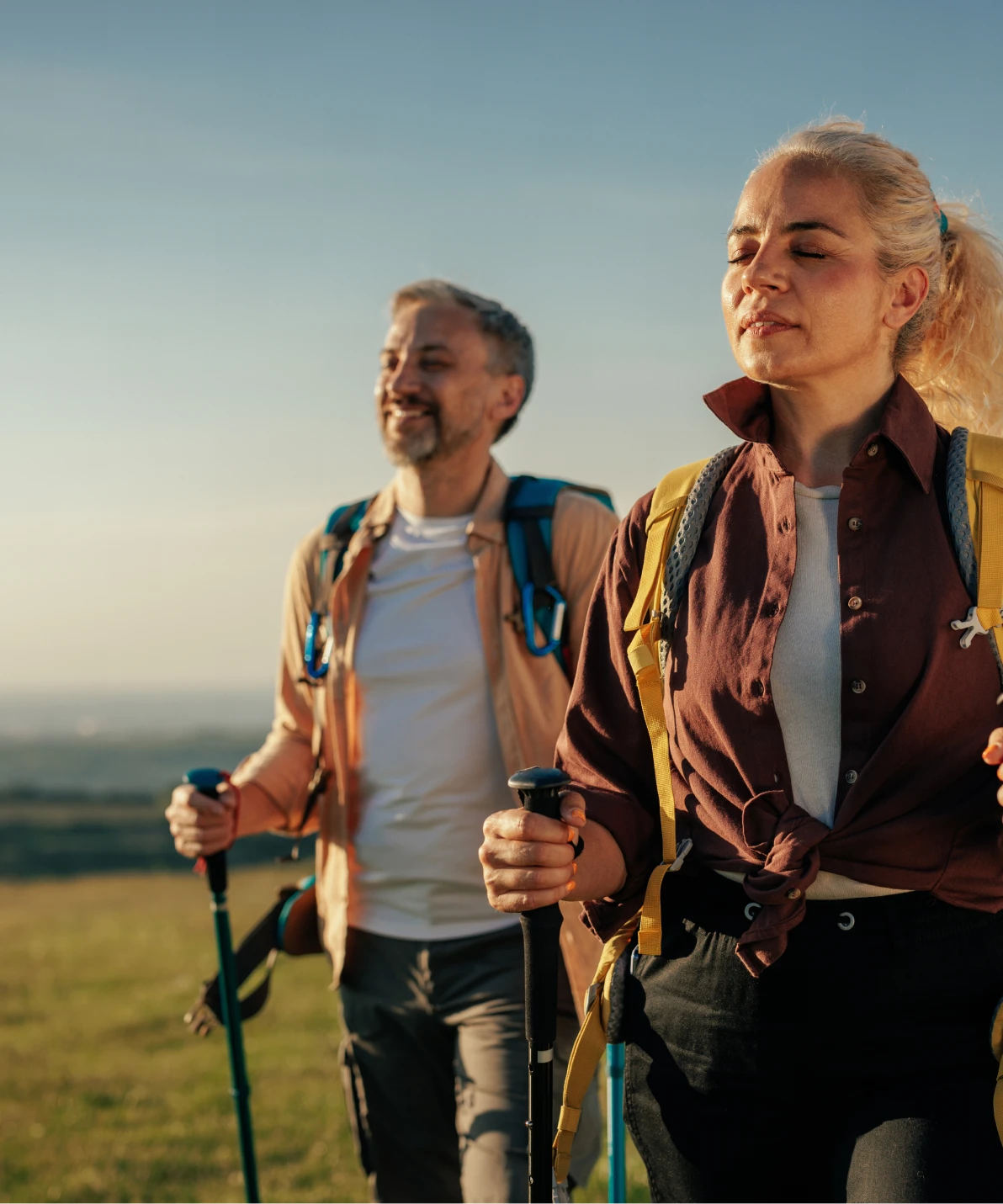 Two people wearing backpacks and holding hiking poles stand on hillside at sunset