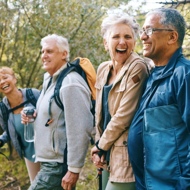 Group of four people wearing backpacks and holding hiking poles stand together on wooded trail, dressed in layers including vests and jackets