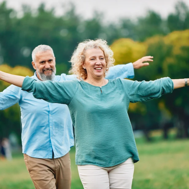 Two people with arms extended outward in outdoor park setting, one wearing teal top and white pants, one wearing light blue shirt and tan pants