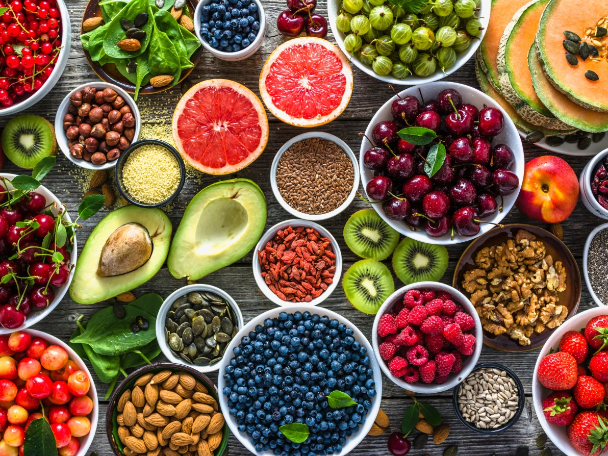 Overhead view of colorful whole foods arranged in bowls including blueberries, raspberries, cherries, avocados, grapefruit, leafy greens, nuts, and seeds