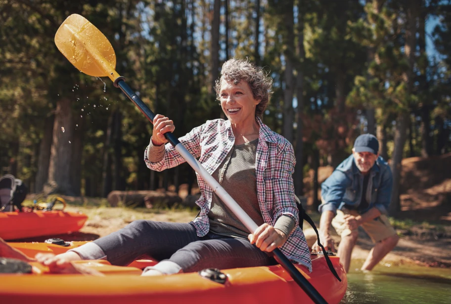 Person in plaid button-up shirt over olive vest sits in orange kayak holding yellow paddle with water droplets, another person behind them