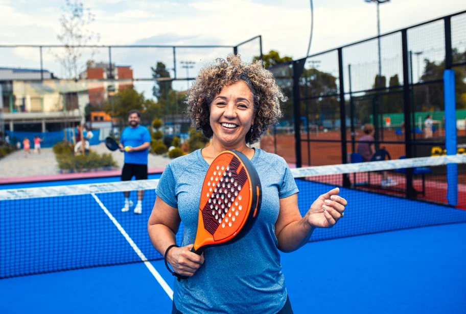 Person with curly hair in light blue athletic top holds orange and blue paddle or racket on bright blue court