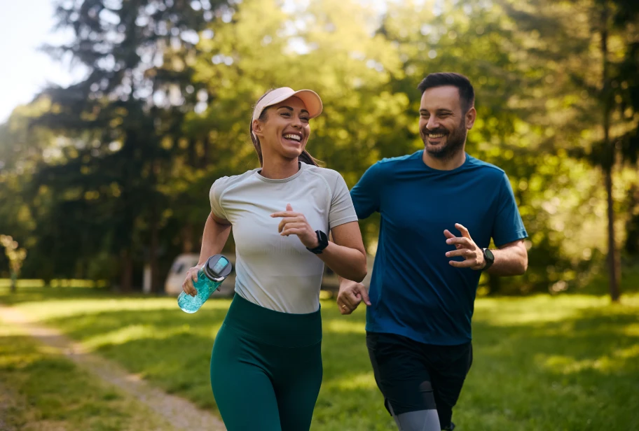 Two people jog side by side on outdoor path, one wearing beige top and one wearing blue top with green shorts