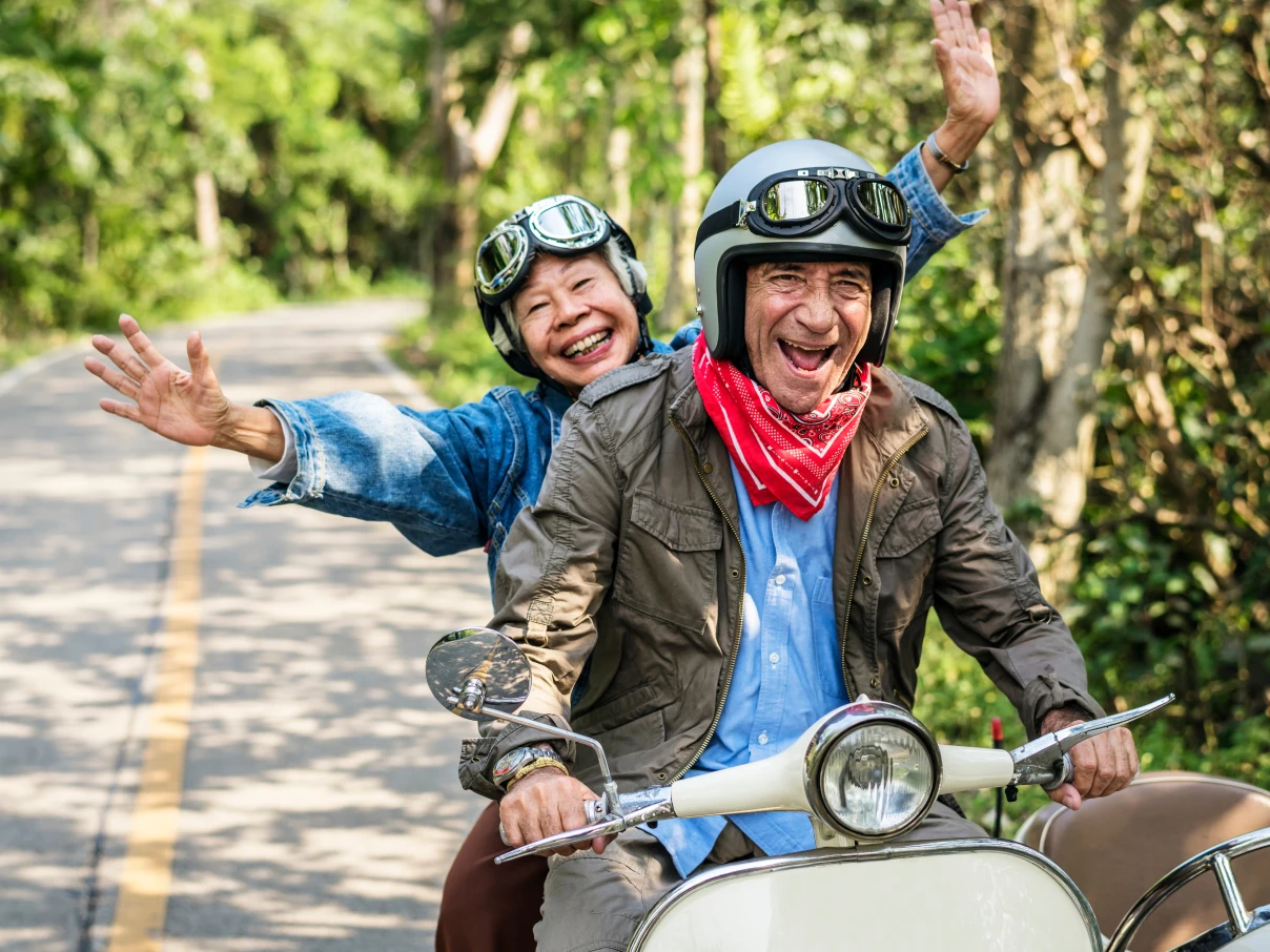 Two people on scooter wearing helmets with goggles on tree-lined road, one in blue denim jacket and one in olive jacket with red bandana