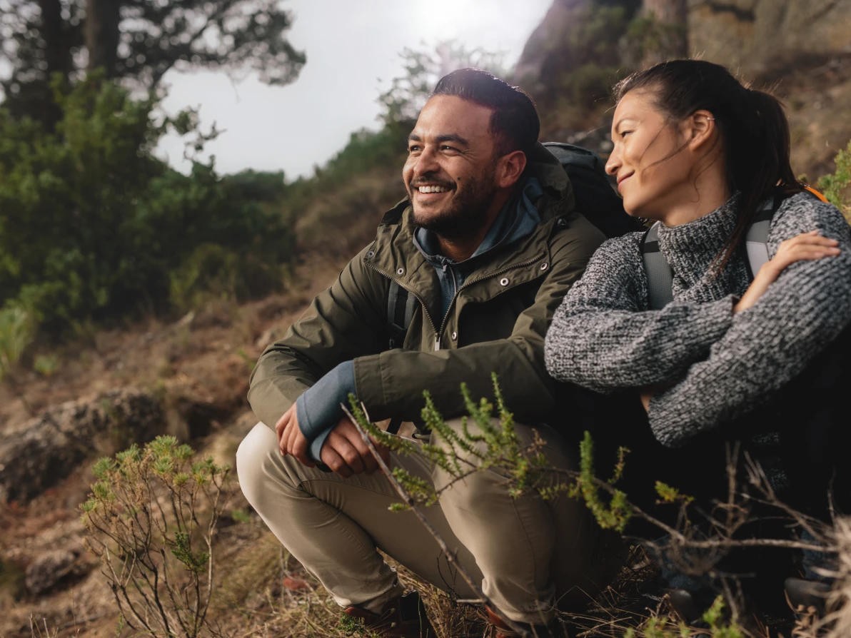 Two people sitting on rocky outdoor terrain with vegetation, one wearing olive green jacket with backpack and one wearing gray knit sweater