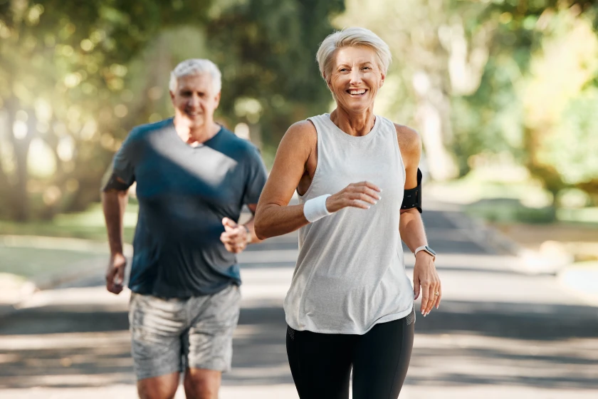 Two people jogging on paved outdoor path, one wearing dark blue top with gray shorts and one wearing white sleeveless top with black pants