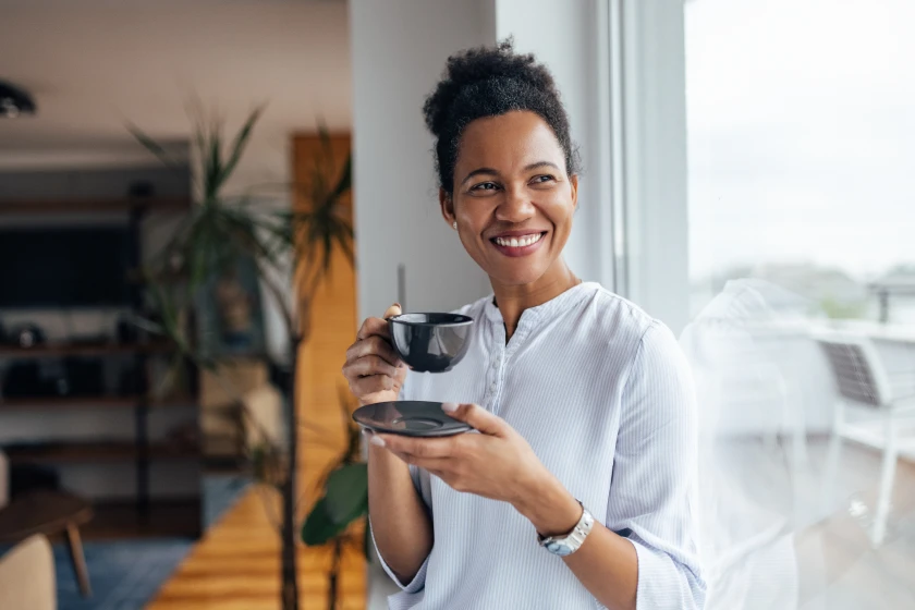 Person in white long-sleeve shirt holds dark coffee cup in bright kitchen with wooden shelving in background