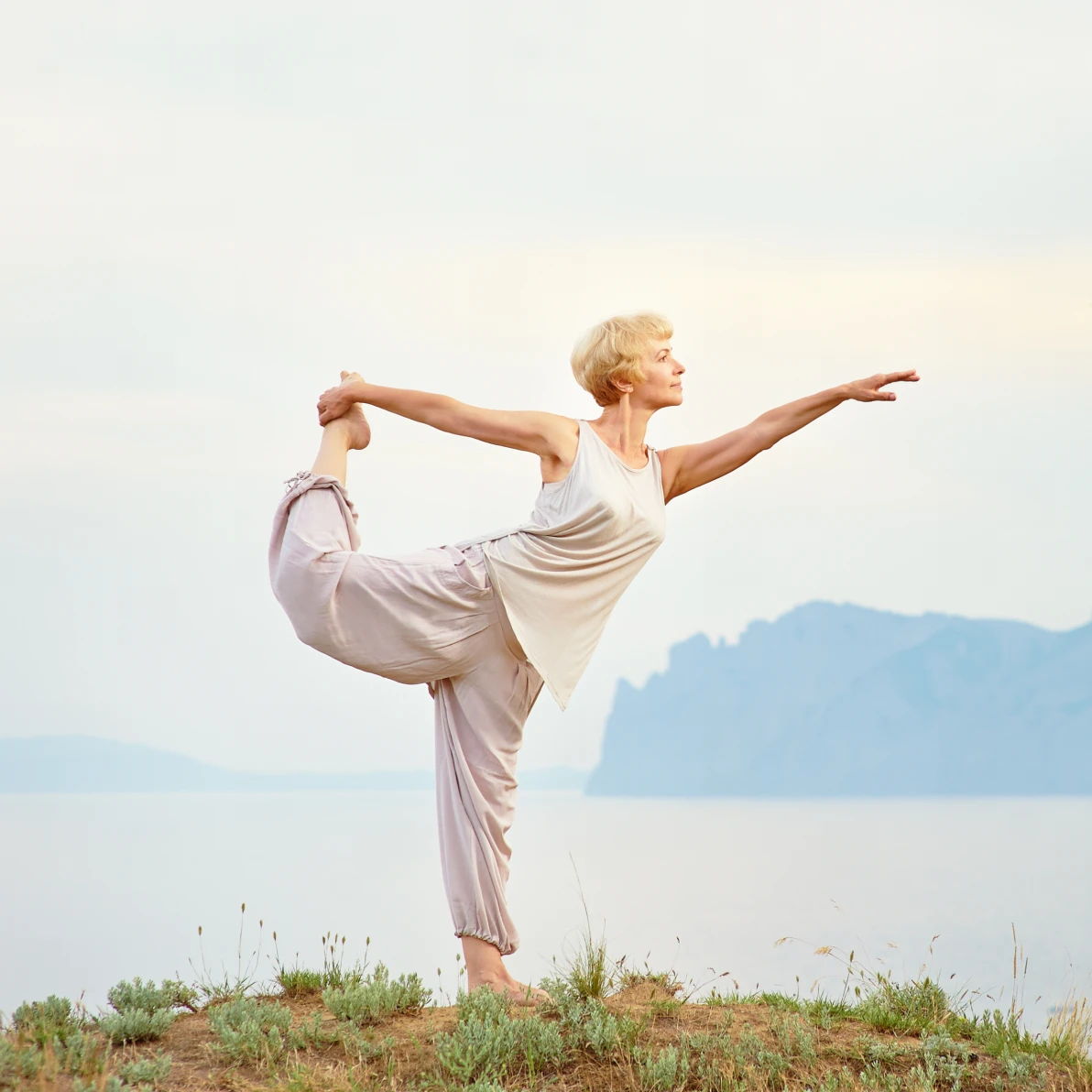 Person in light-colored clothing standing on one leg with arms extended outdoors near body of water